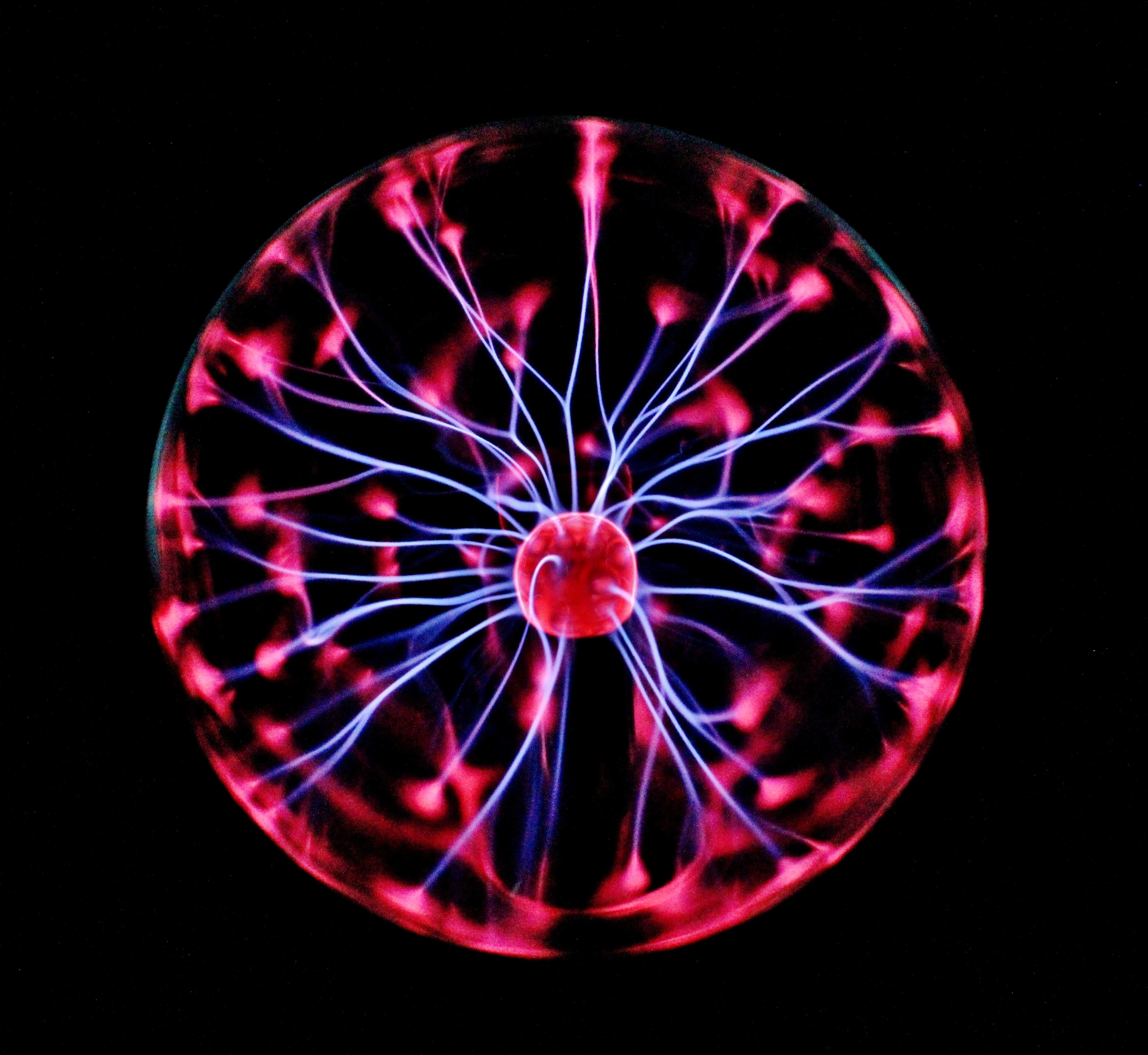 Plasma ball, a clear spherical glass container that is filled with a low-pressure mixture of gases that are ionized by an electric current, creating colorful streamers of light within. A bright purple ball is at the center of the image, with the light streaming out in tendrils of white, blue and pink. The tendrils appear to be flowing out of the plasma ball and they are not symmetrically arranged. The background is completely black, with no visible shapes or textures.