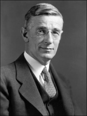 A man who appears to be in his fifties or sixties, with short hair and a serious expression on his face. He is wearing a suit and tie, and is sitting at a desk with papers and a telephone on it. The man is identified by a caption below the photograph as Vannevar Bush.
